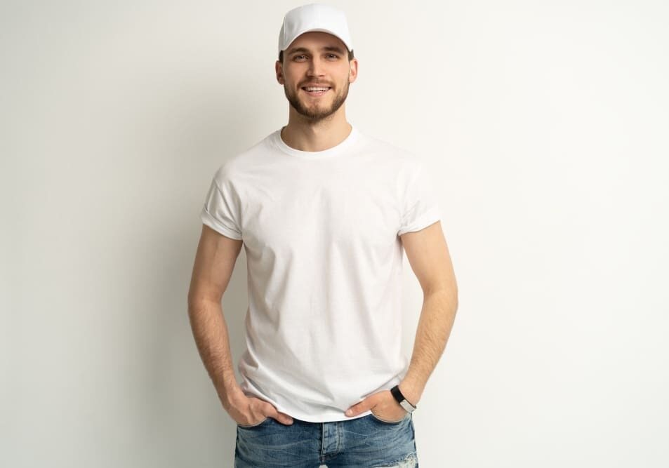 A man wearing white shirt and hat standing next to wall.